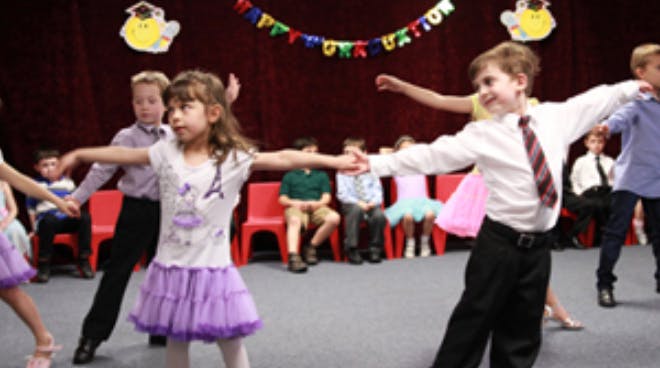 Children dancing in a room with other children seated in the background