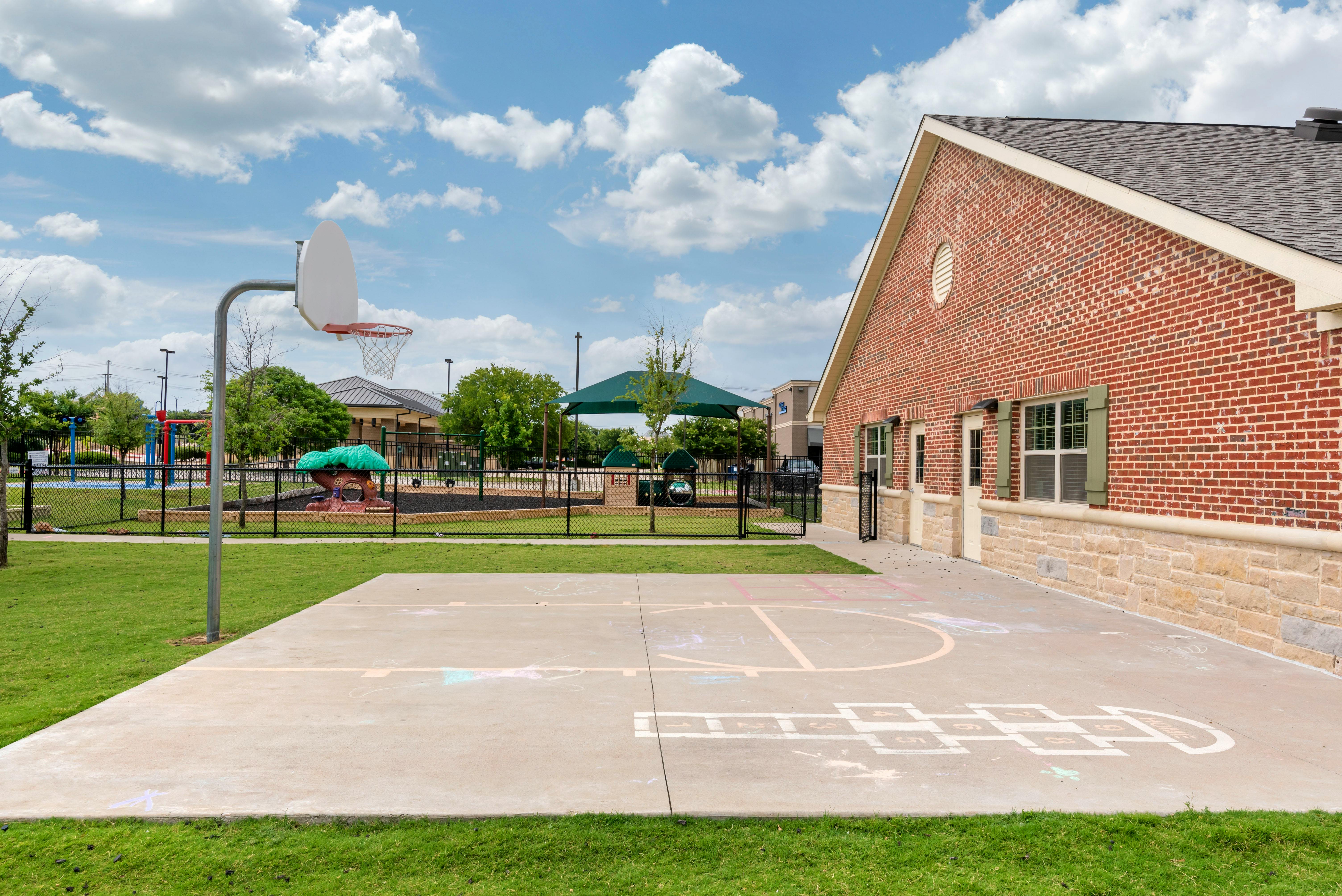 Basketball hoop on a paved court next to a red brick building and fenced playground