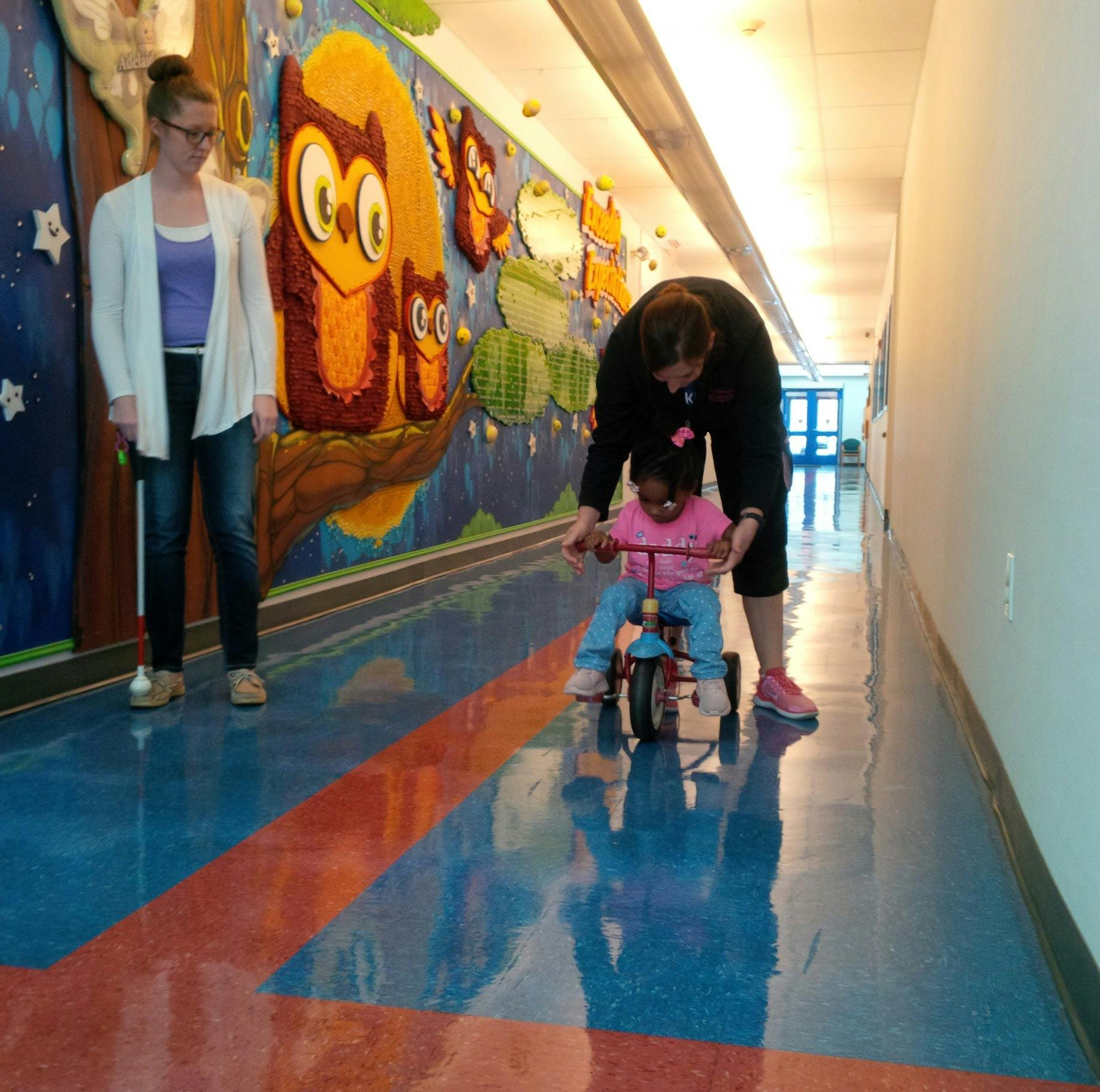 Person helping a child on a tricycle in a hallway with a mural