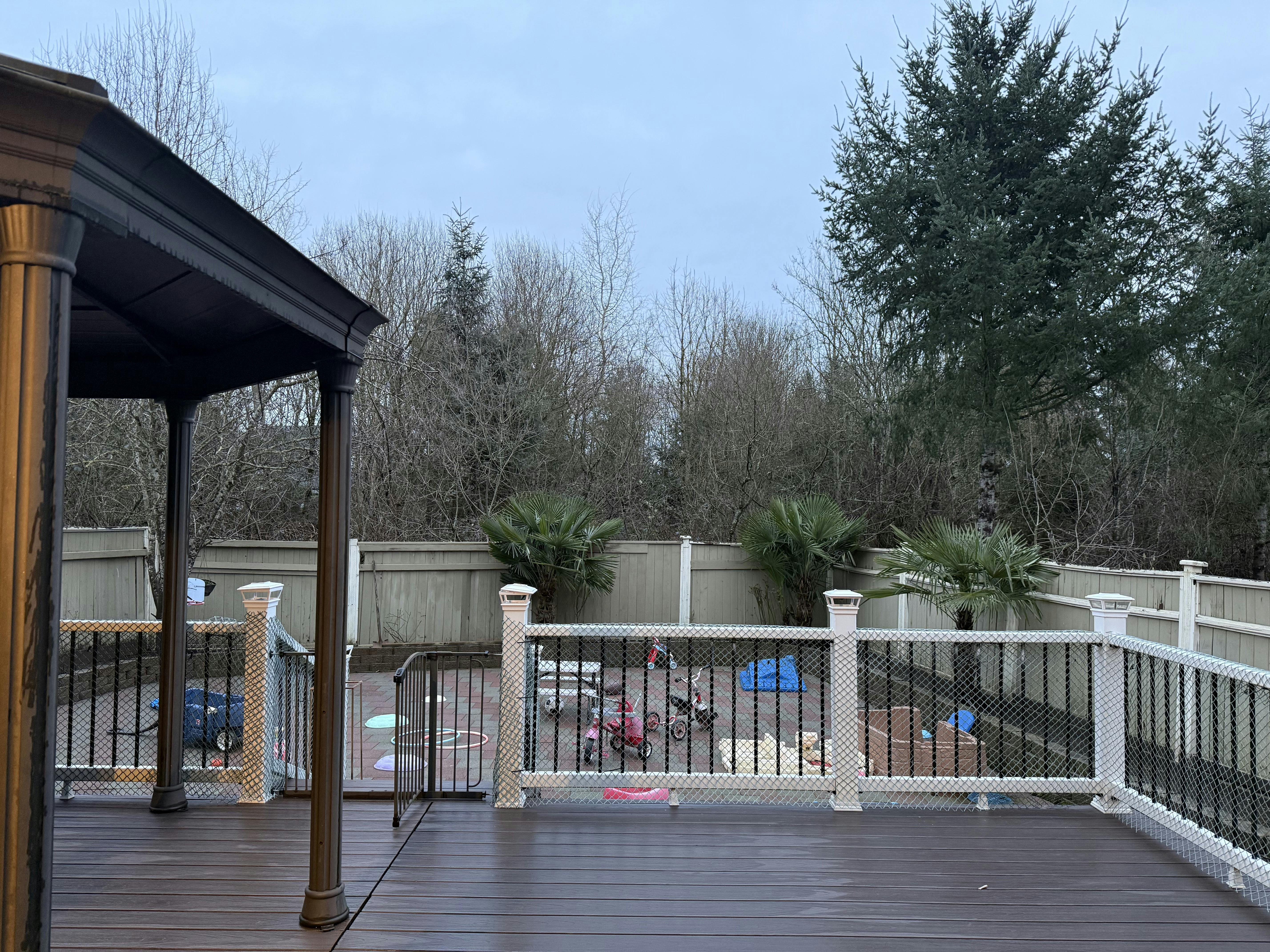 A wooden deck overlooking a fenced yard with toys, trees, and palm trees
