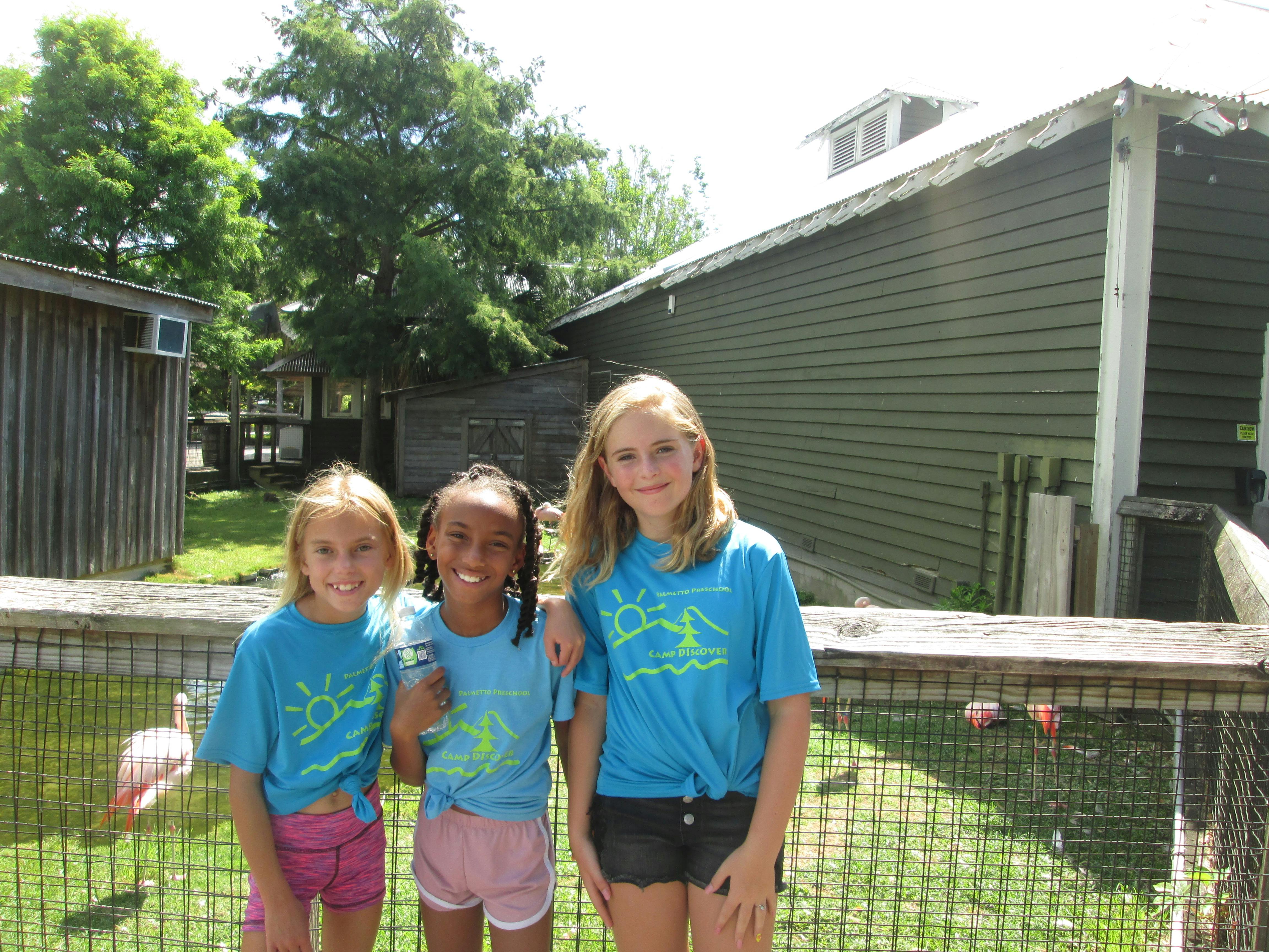 Three children in blue Camp Discover shirts standing next to a fence