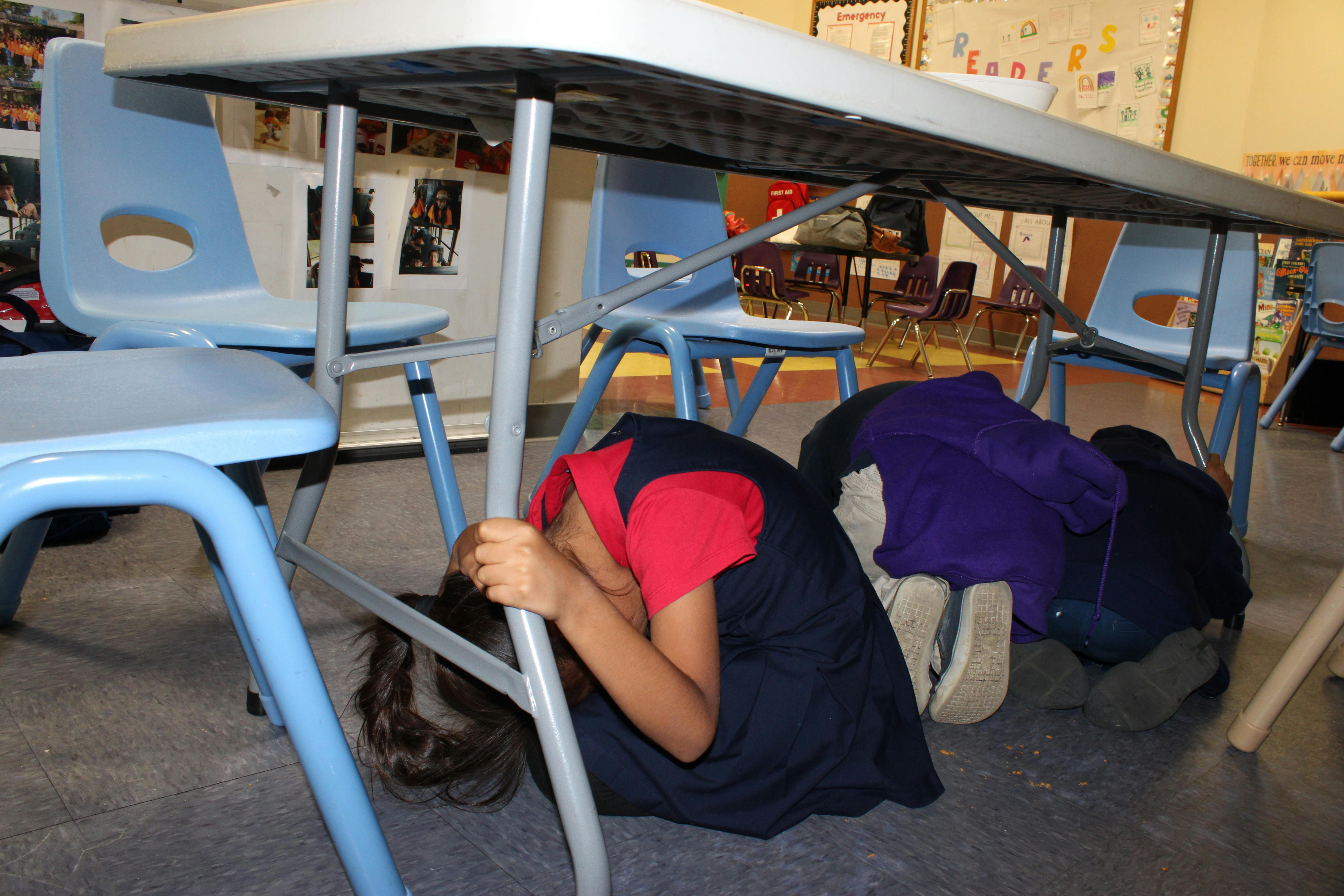children and teens crouching under a table in a classroom