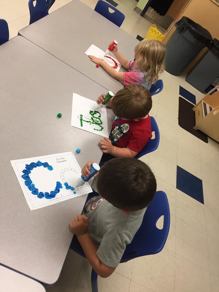 Three children sitting at a table doing an art activity with dot markers