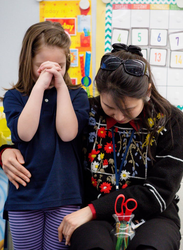 Child standing with hands clasped, person sitting nearby with hands on the child's back
