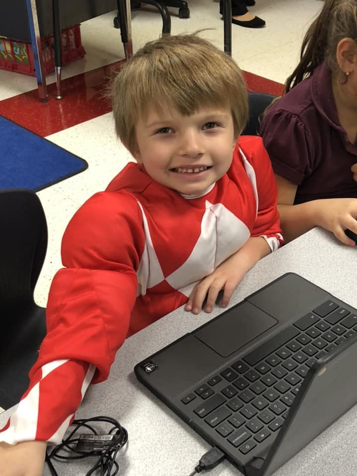 Child in red and white shirt sitting at a desk with a laptop