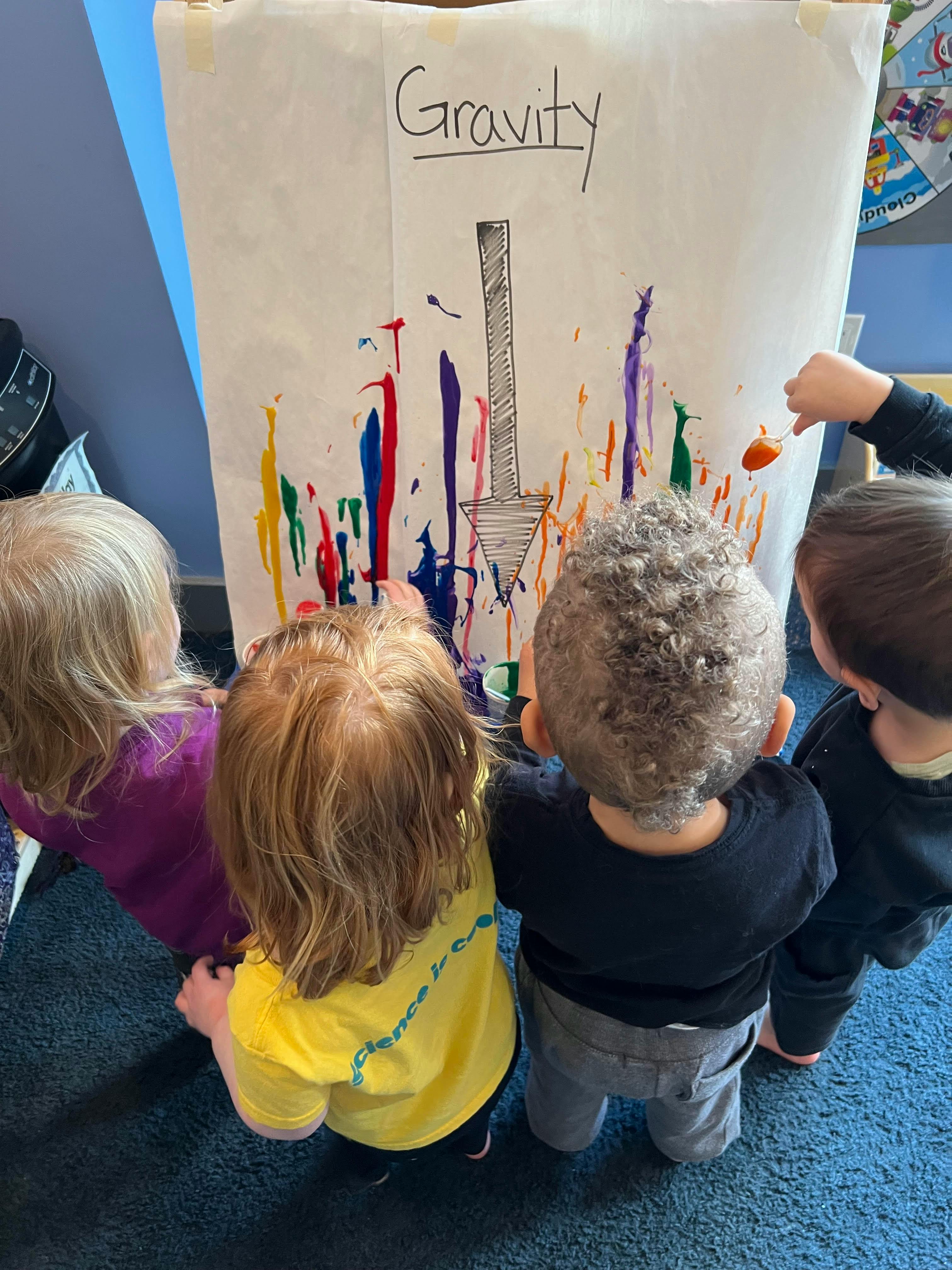 Children painting on a large sheet of paper with "Gravity" written at the top