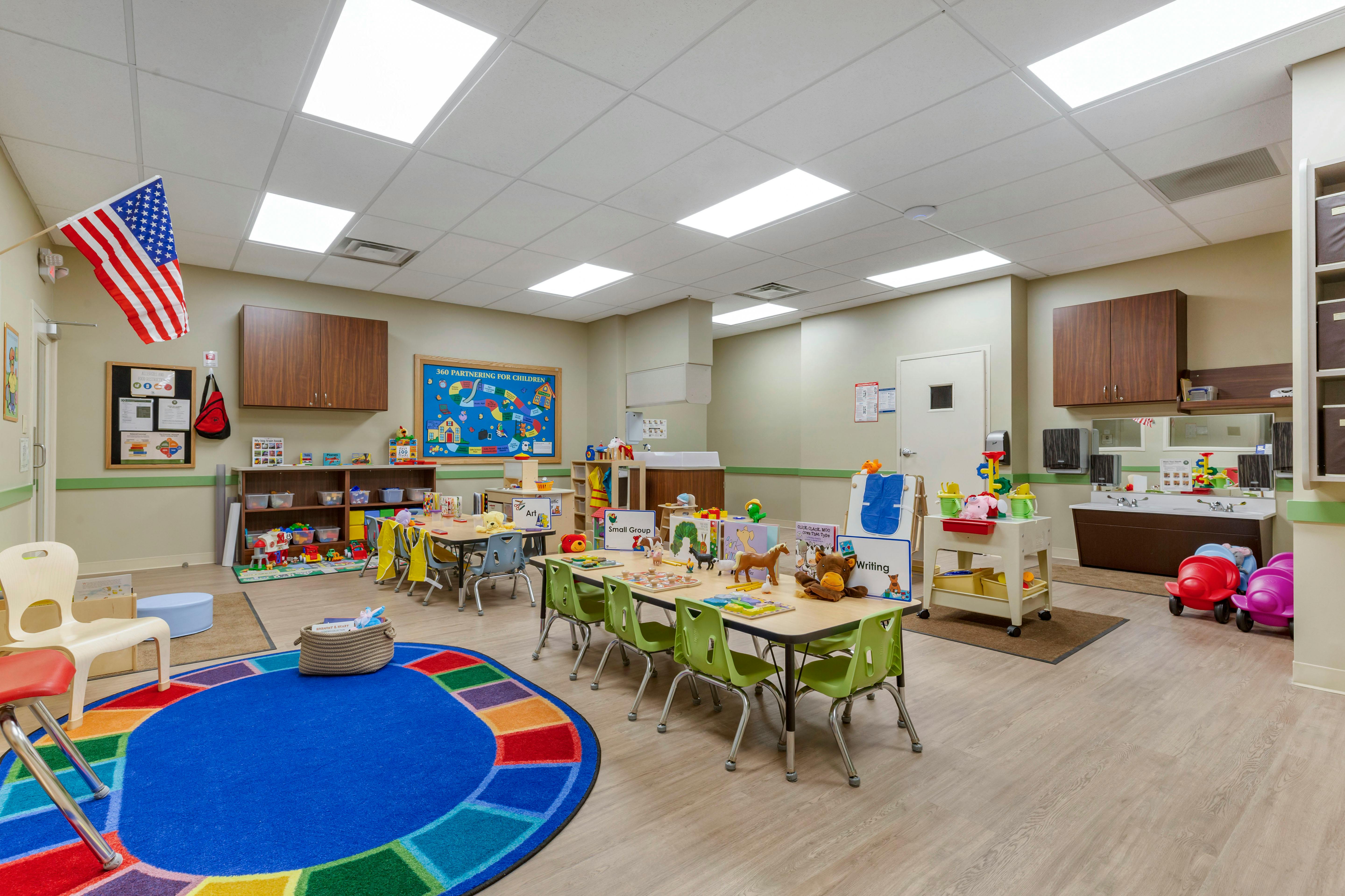 A colorful classroom with small tables and chairs, a play area, and a sink