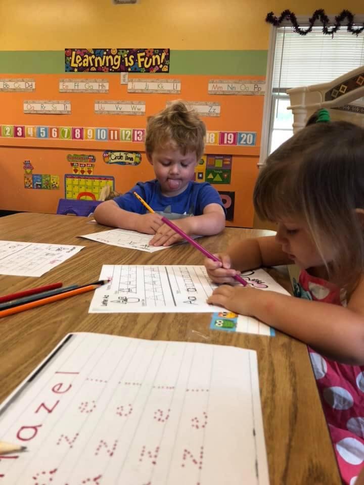 Two children writing at a table in a classroom with "Learning is Fun!" sign