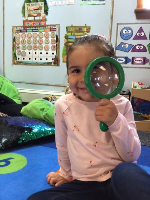 Child holding a magnifying glass over one eye in a classroom