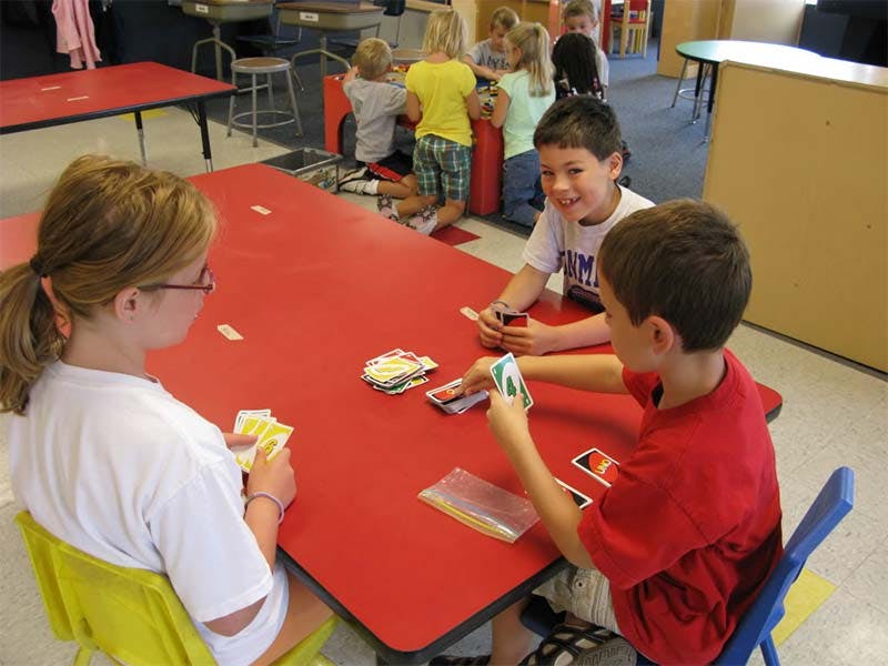 Three children playing cards at a red table in a classroom