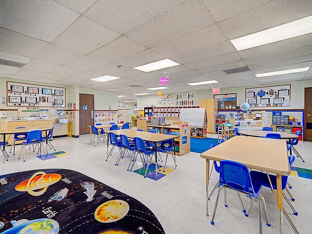 Classroom with multiple tables and blue chairs, space-themed rug in foreground