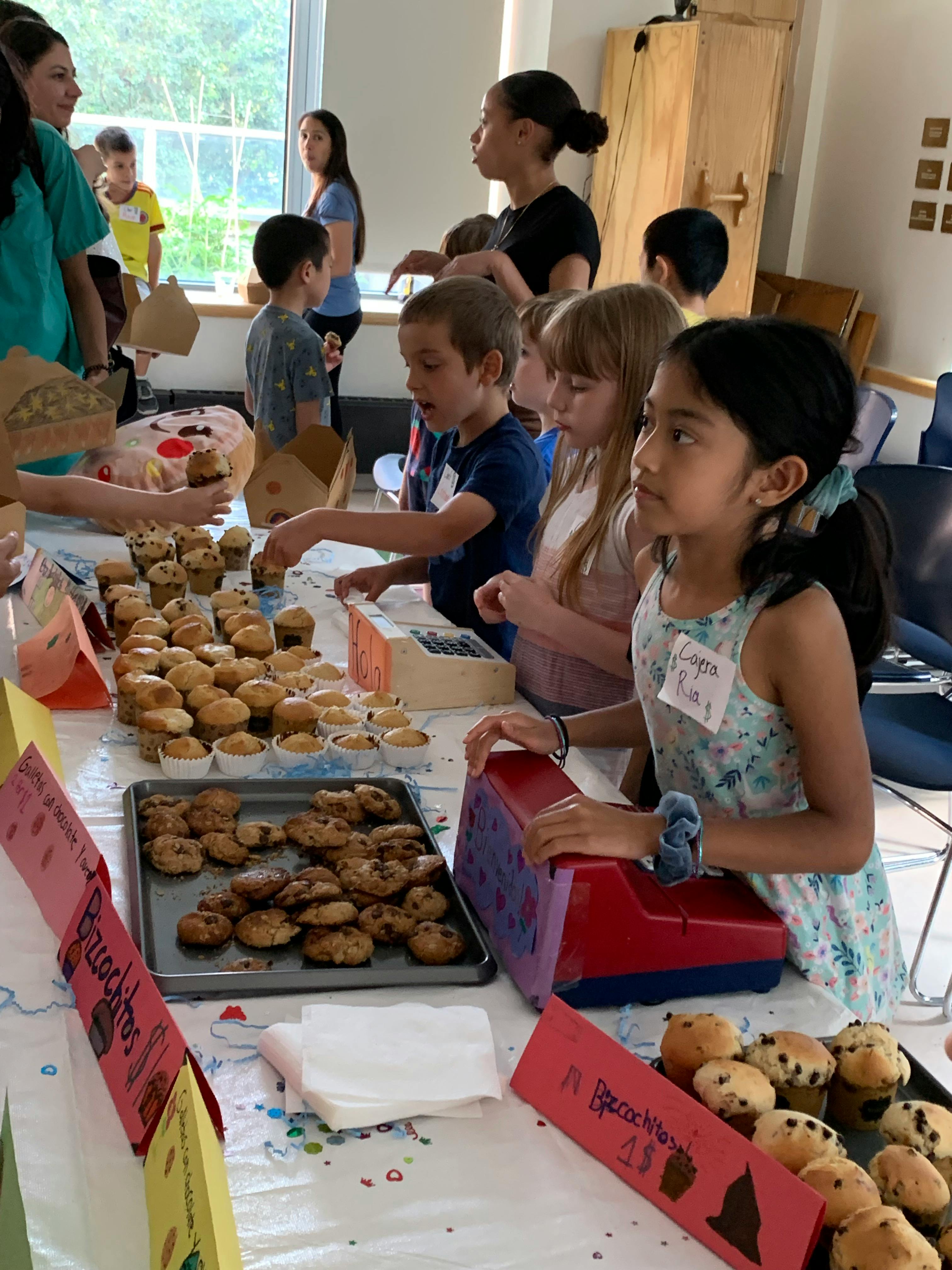 Children working at a bake sale table with trays of muffins and signs for bizcochitos