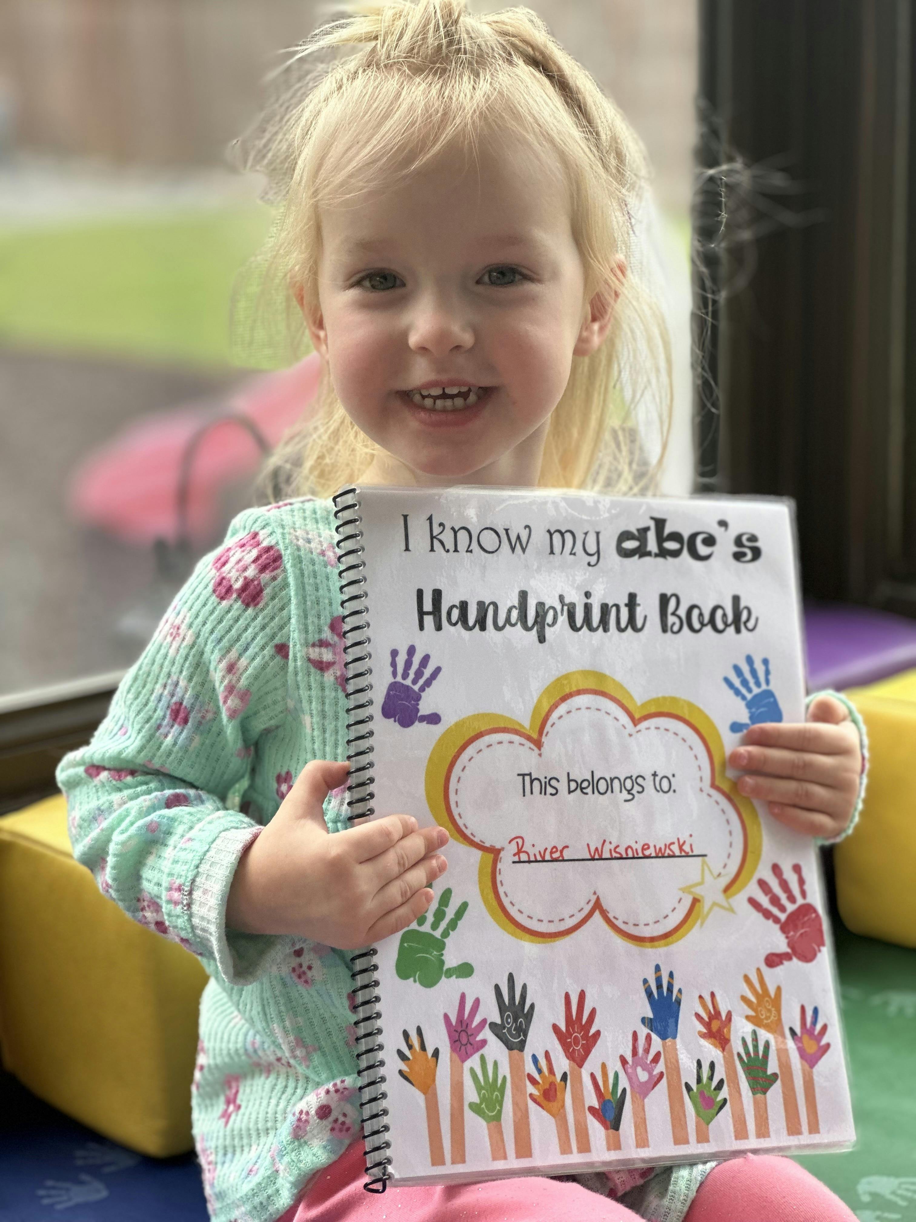 Child holding a book titled "I know my abc's Handprint Book"