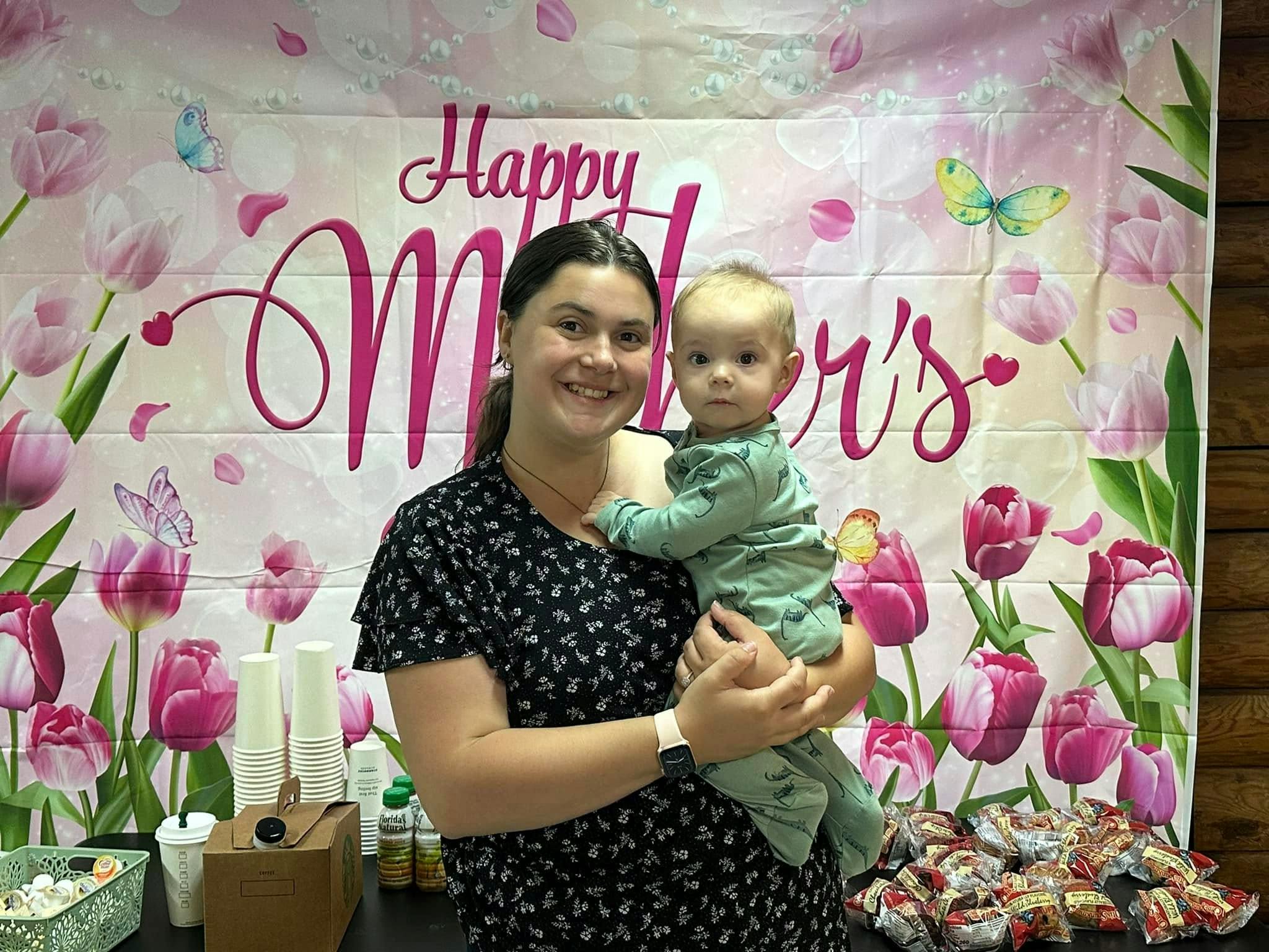 Person holding a child in front of a banner that says "Happy Mother's"