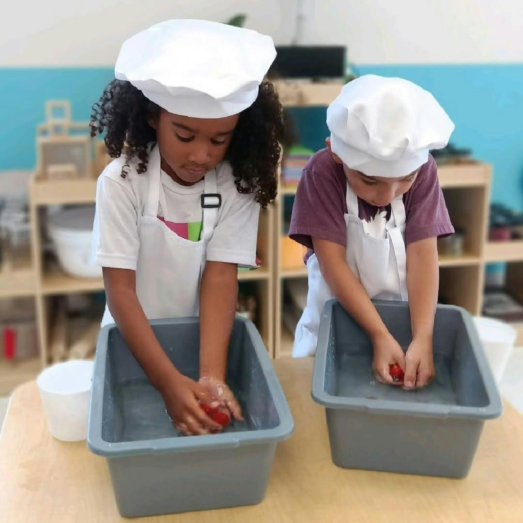 Two children in chef hats and aprons washing items in bins