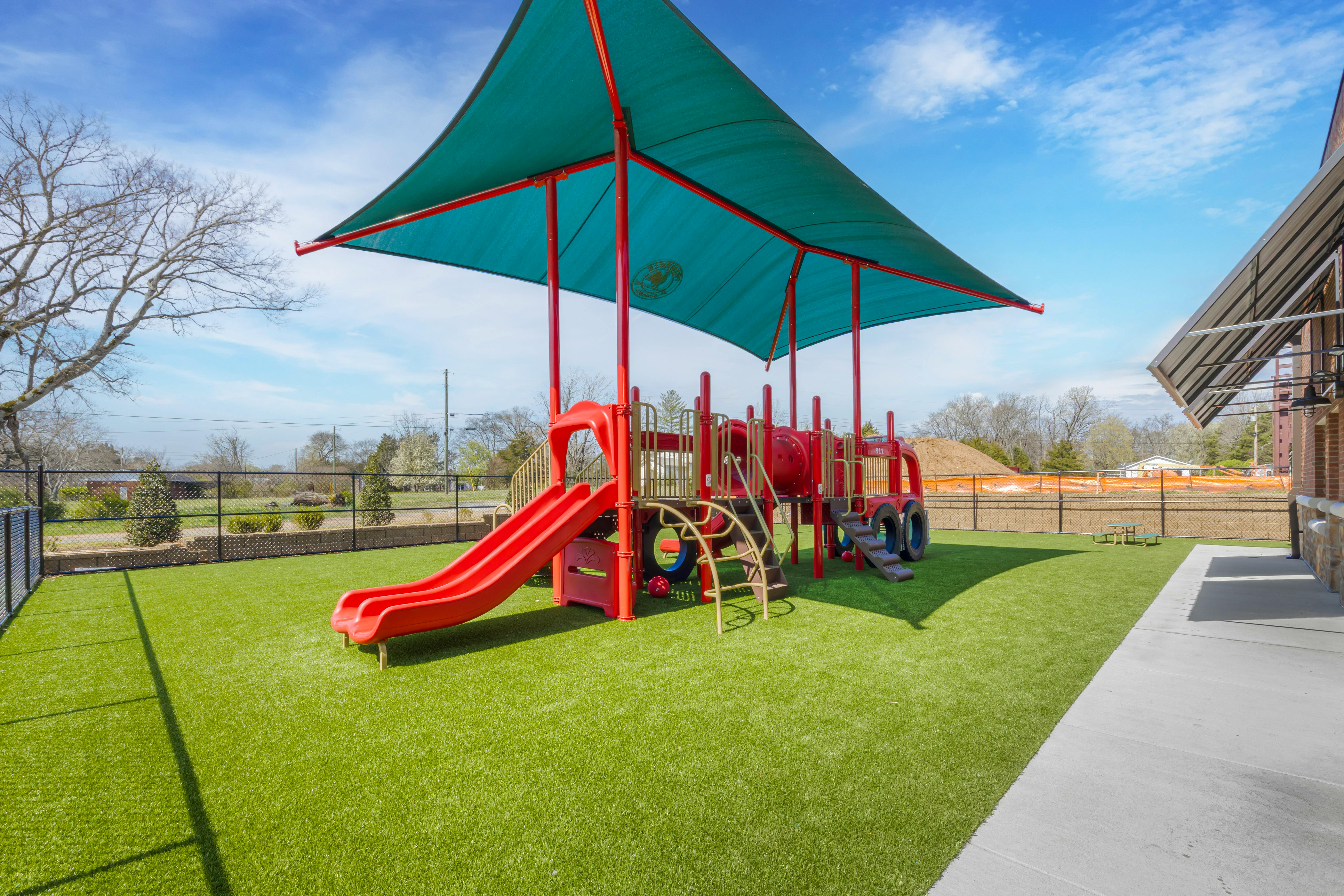 Red playground structure with slides and climbing elements on green turf under a shade