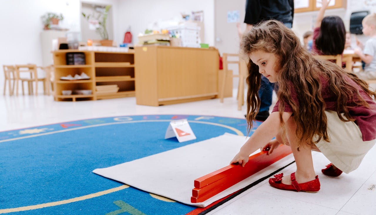 Child crouching and arranging red blocks on a white mat in a classroom