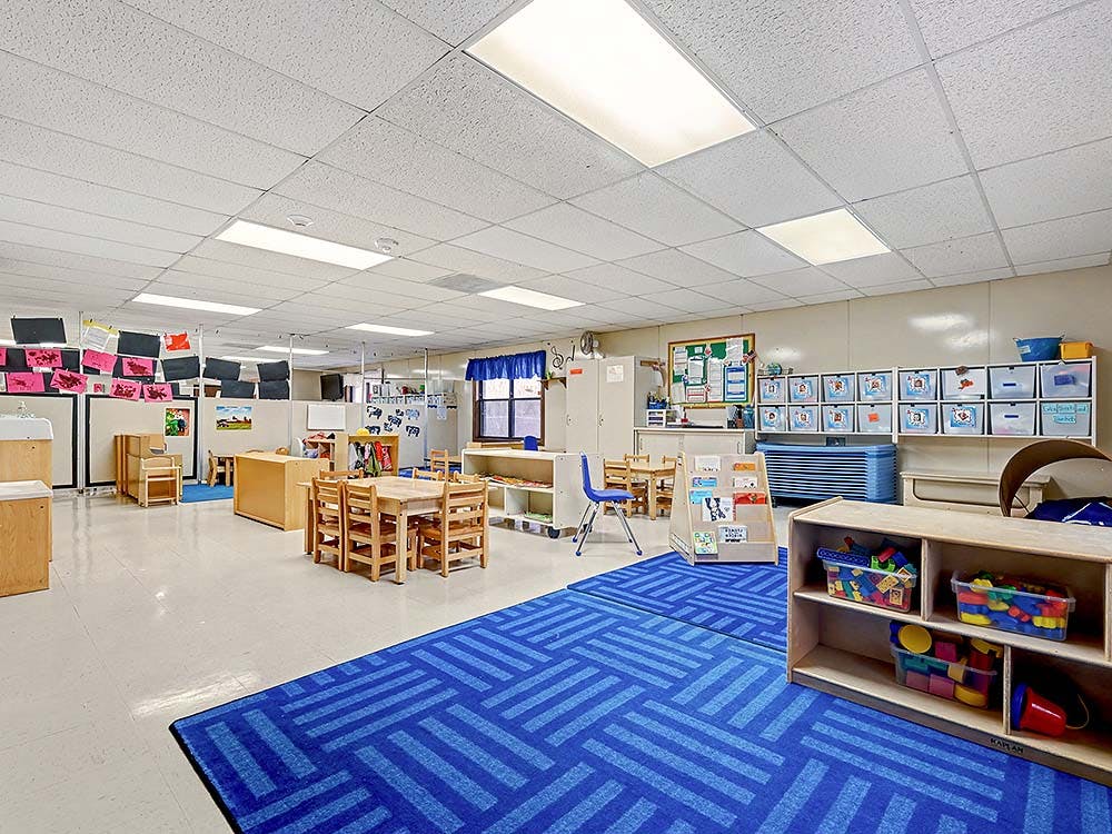 Empty classroom with small wooden tables and chairs, a blue patterned rug, and cubbies