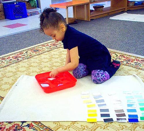 Child kneeling on a rug, sorting small items from a red tray onto a white mat