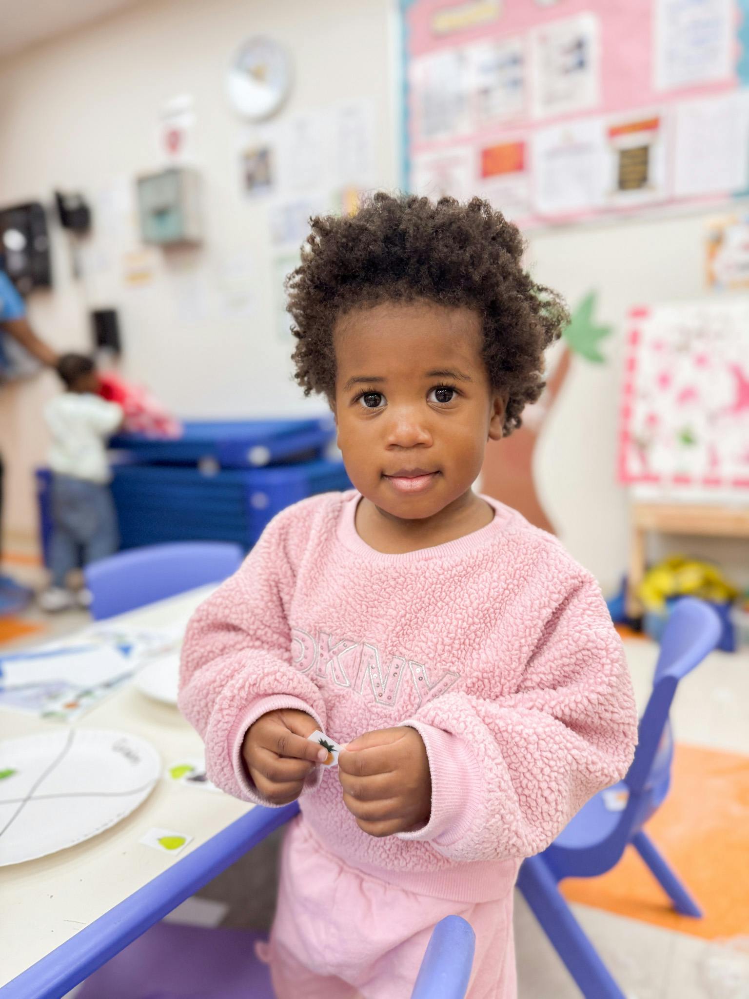 Child in a pink sweater holding a small object in a classroom