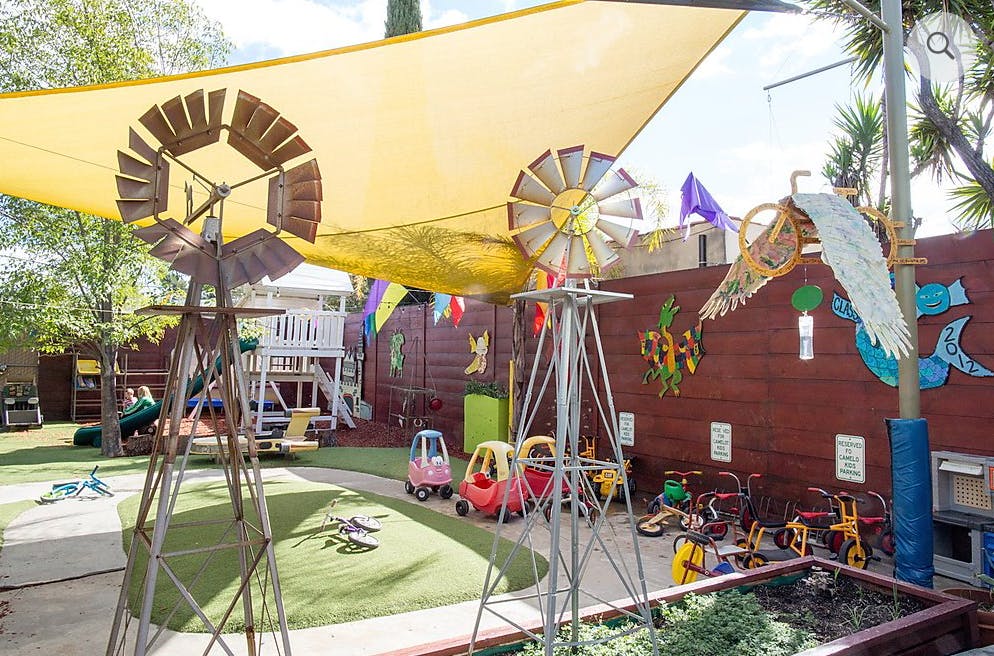 Outdoor play area with shade sail, two decorative windmills, and ride-on toys