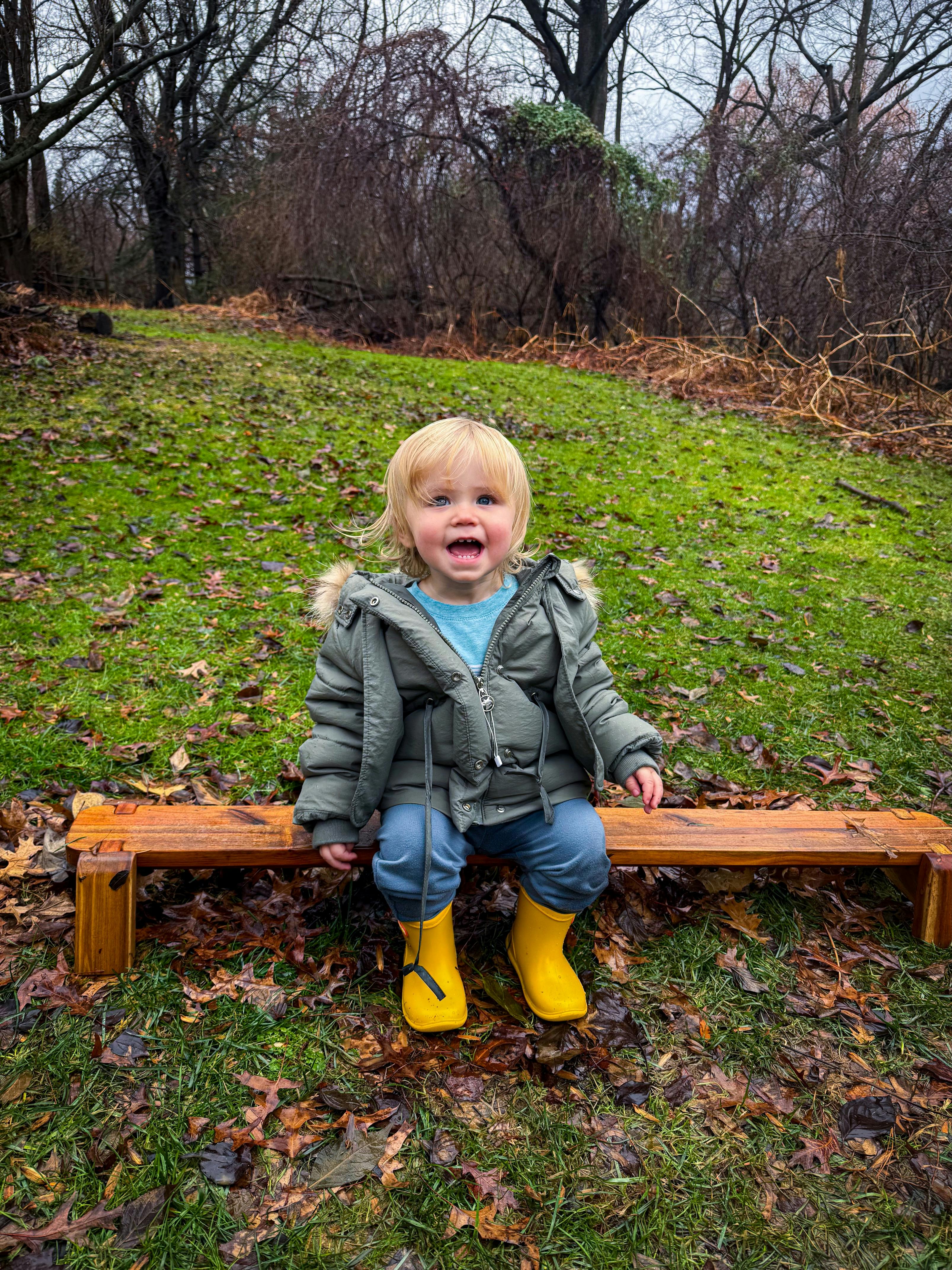Child sitting on a wooden bench in a park wearing yellow rain boots