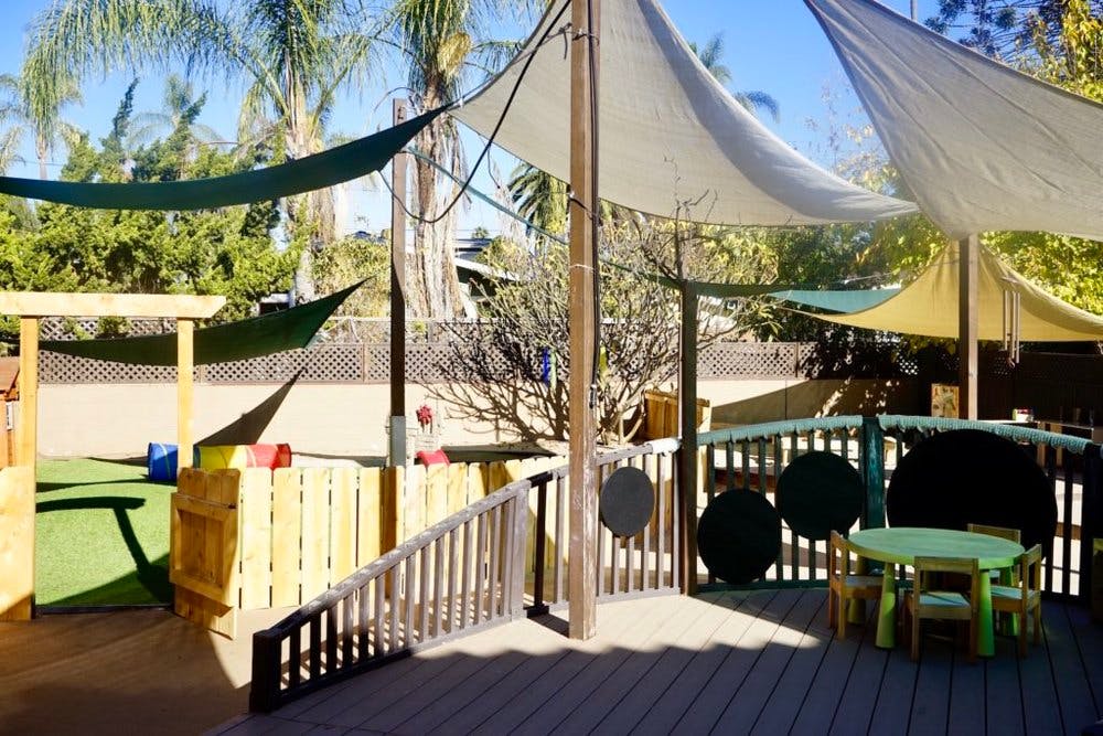 Outdoor play area with shade sails, wooden fence, and small table with chairs