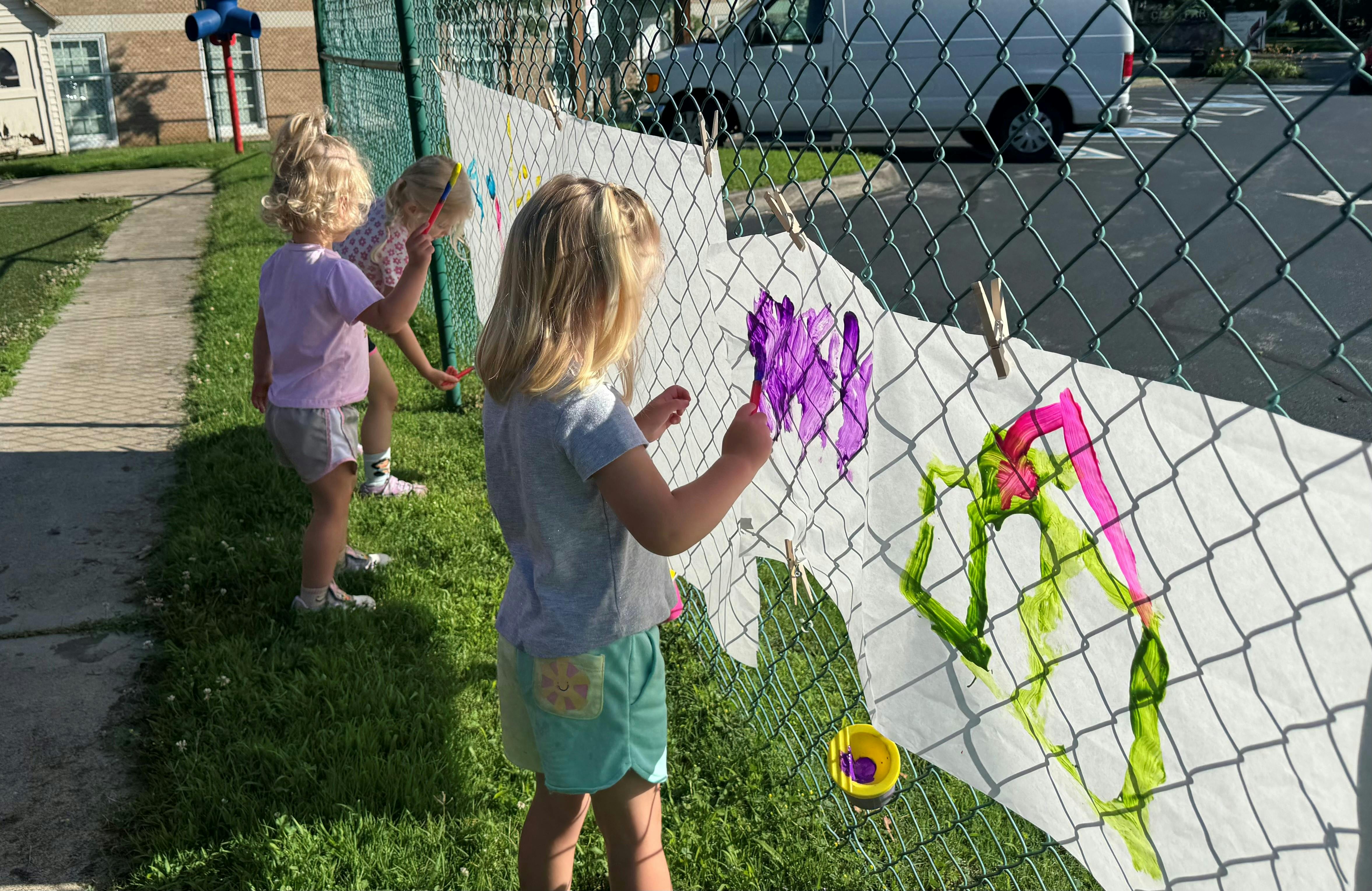 Three children painting on paper attached to a chain-link fence
