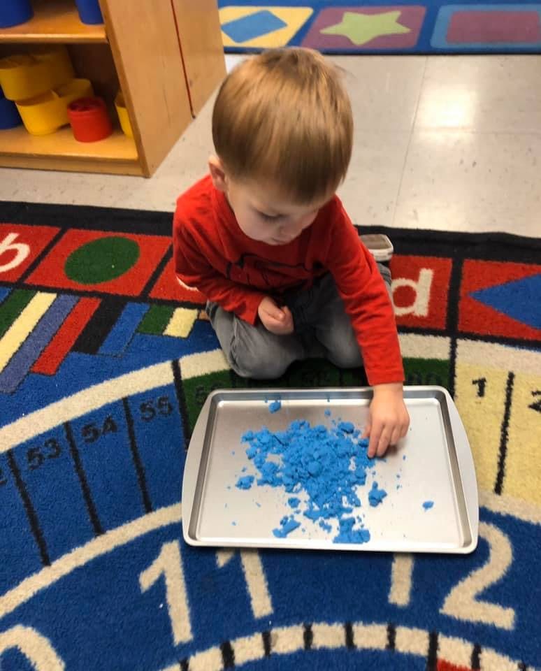 Child kneeling on a rug, playing with blue kinetic sand on a tray