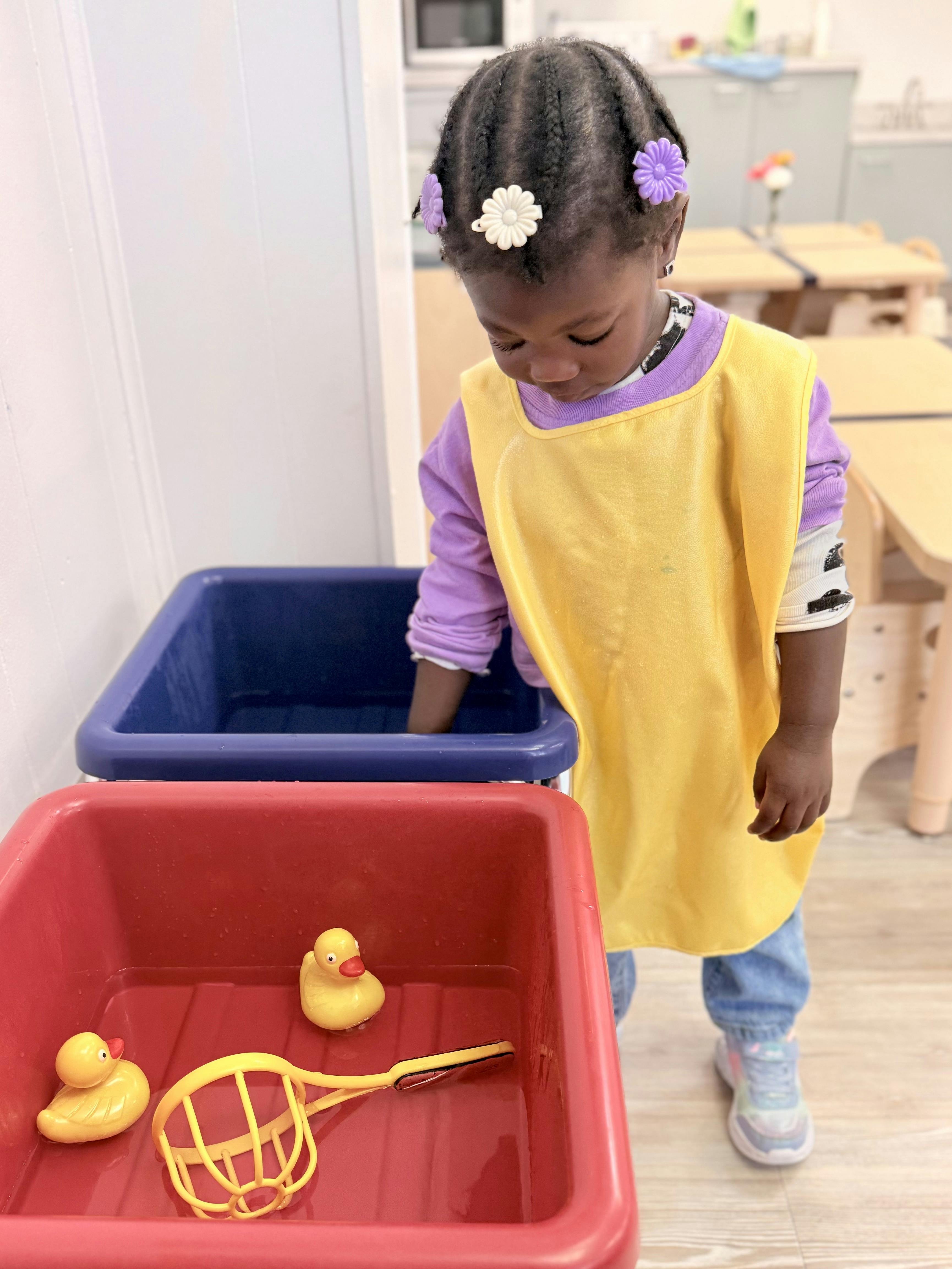 Child in yellow smock playing with rubber ducks and scoop in water bins