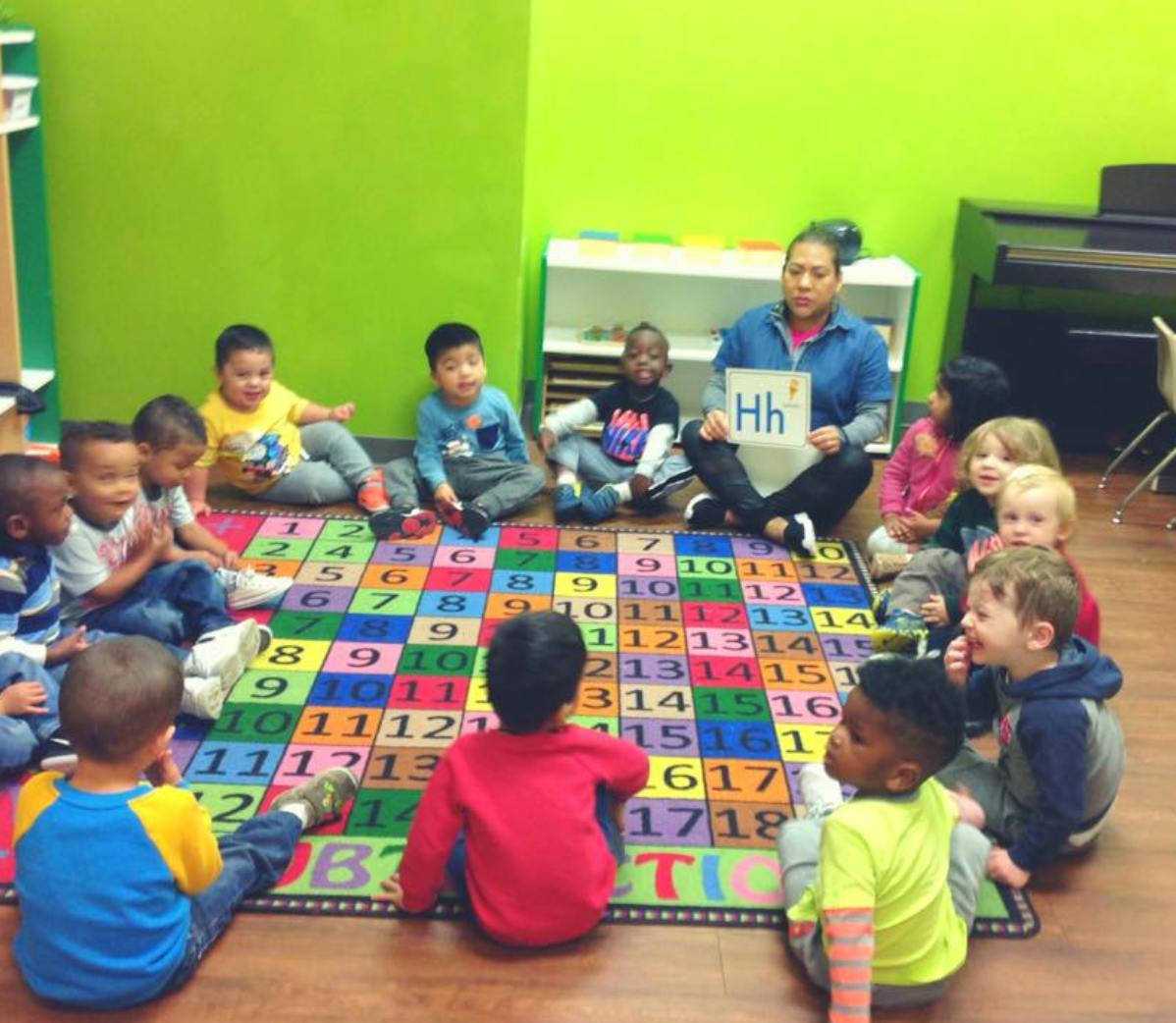 Adult holding up a letter H sign to a group of children sitting on a number rug