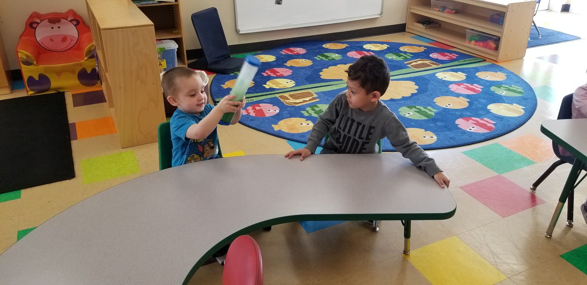 Two children at a table in a classroom, one holding up a toy