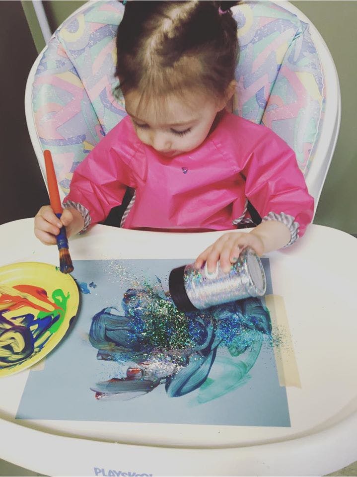 Child in a high chair painting and pouring glitter on paper