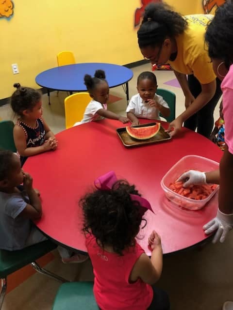 Adults serving watermelon to children at a red table