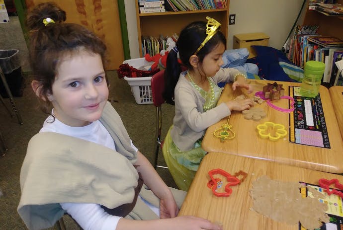 Child in costume sitting at a desk, another child playing with dough