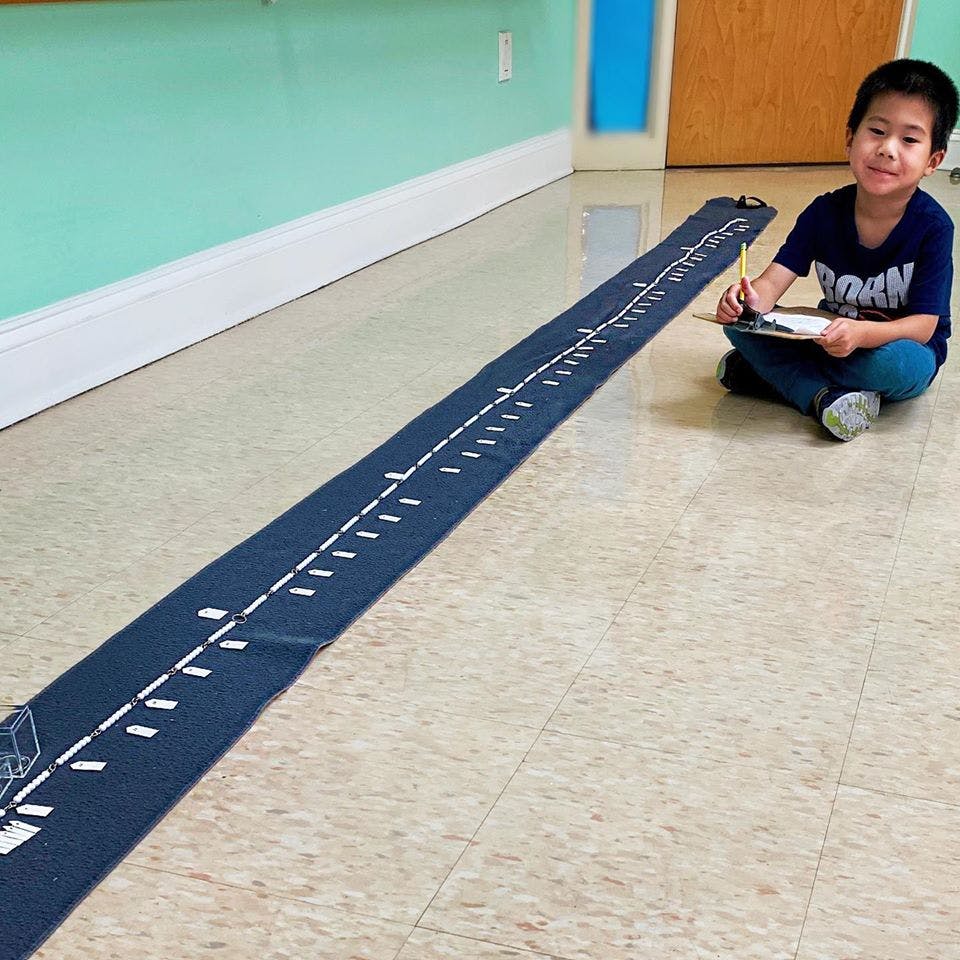 Child sitting on floor next to a long number line, holding a clipboard and pencil