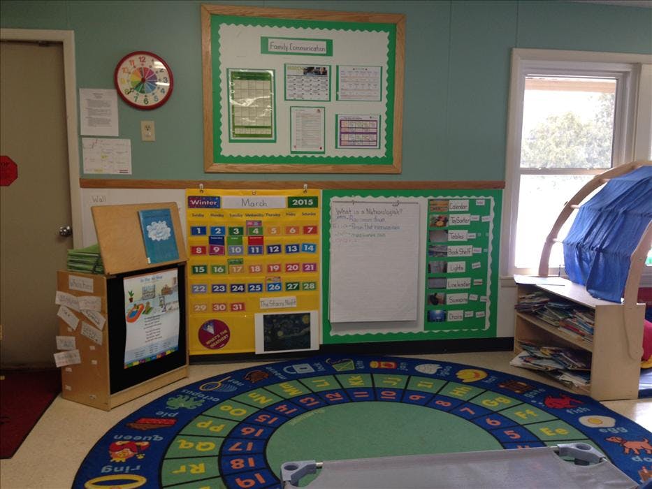 A classroom corner with a calendar, whiteboard, colorful rug, and child's furniture