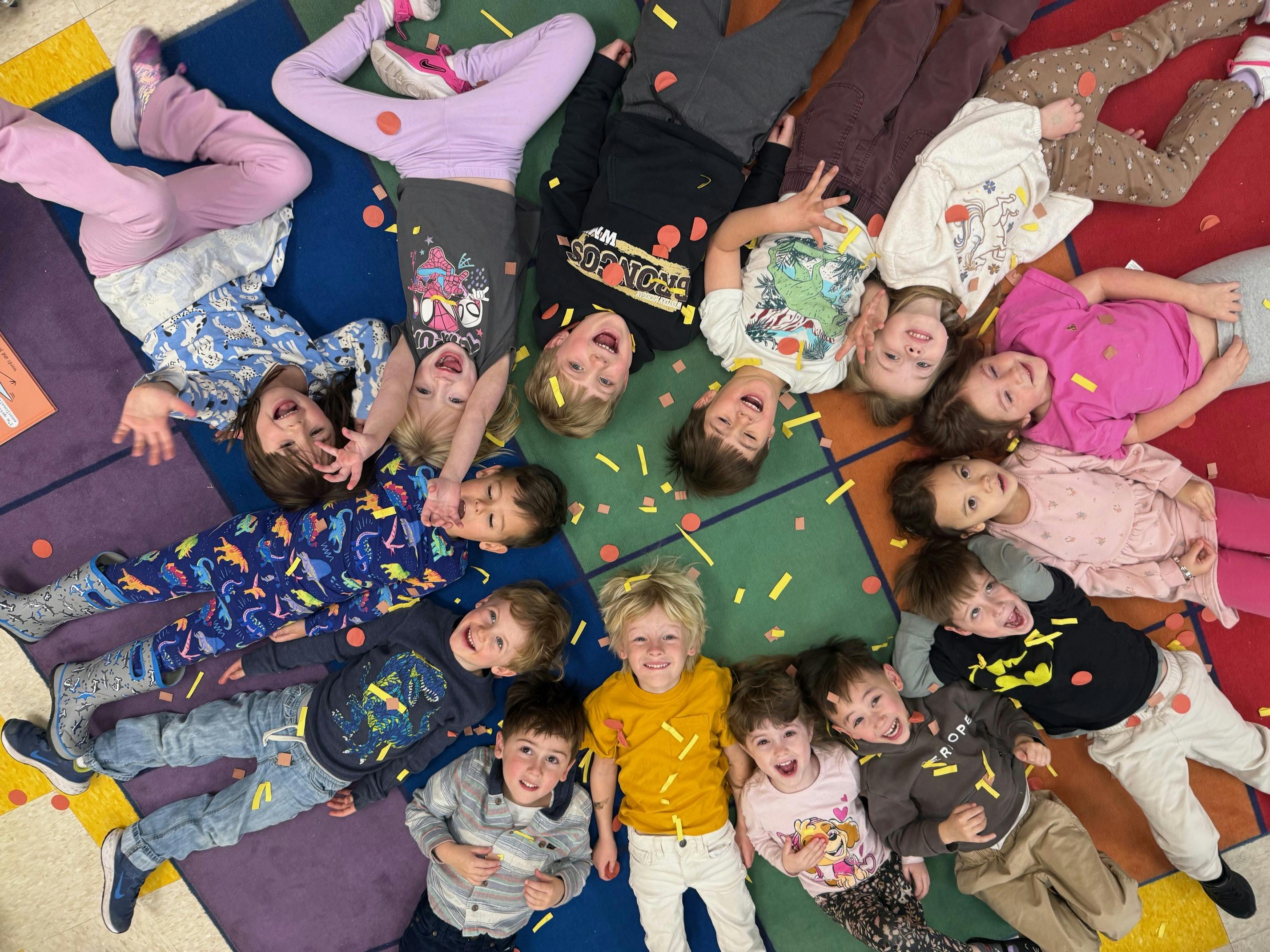 Group of children lying on a colorful rug covered in confetti, looking up and smiling
