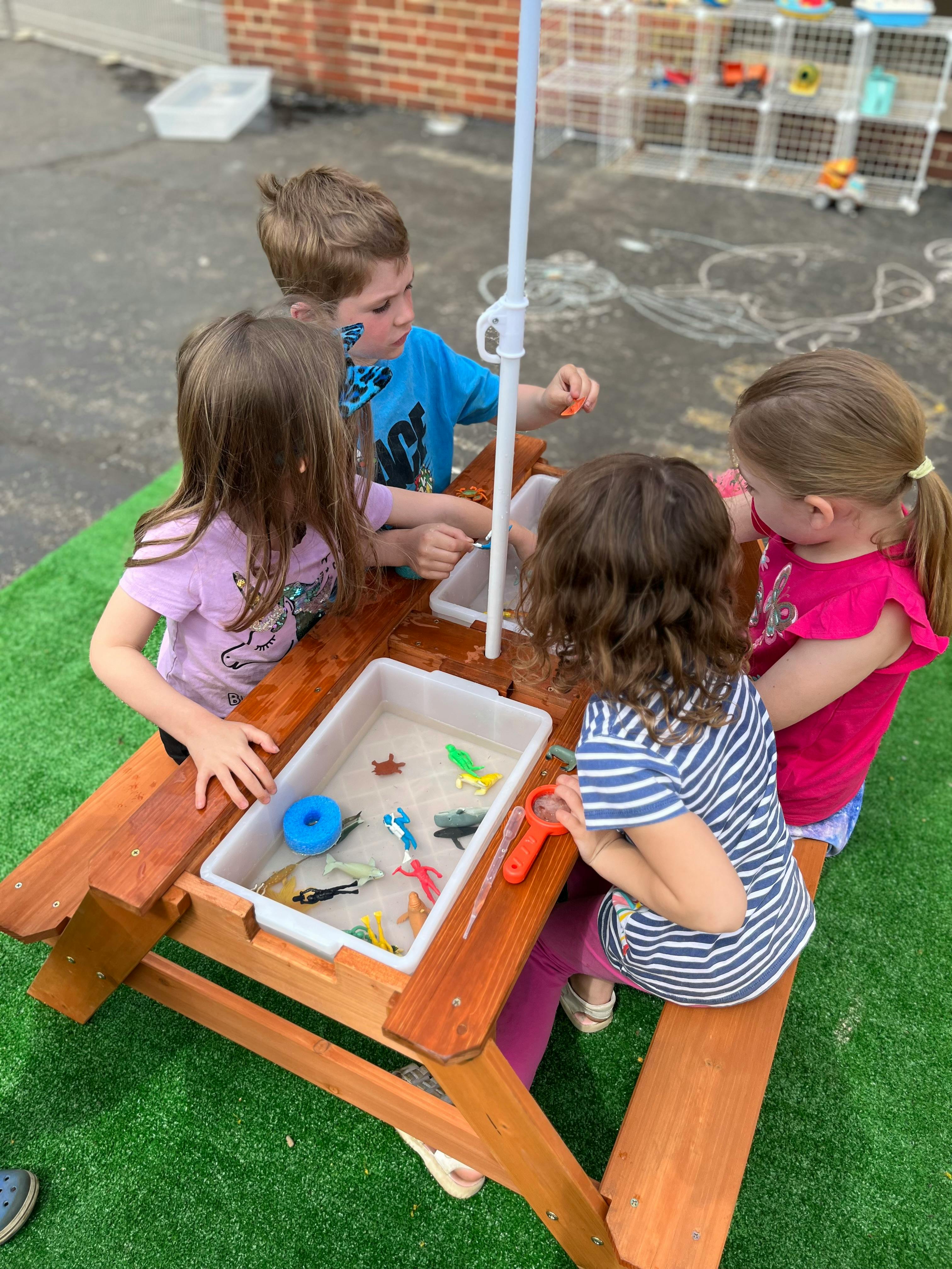 Four children playing at a wooden picnic table with a sensory bin