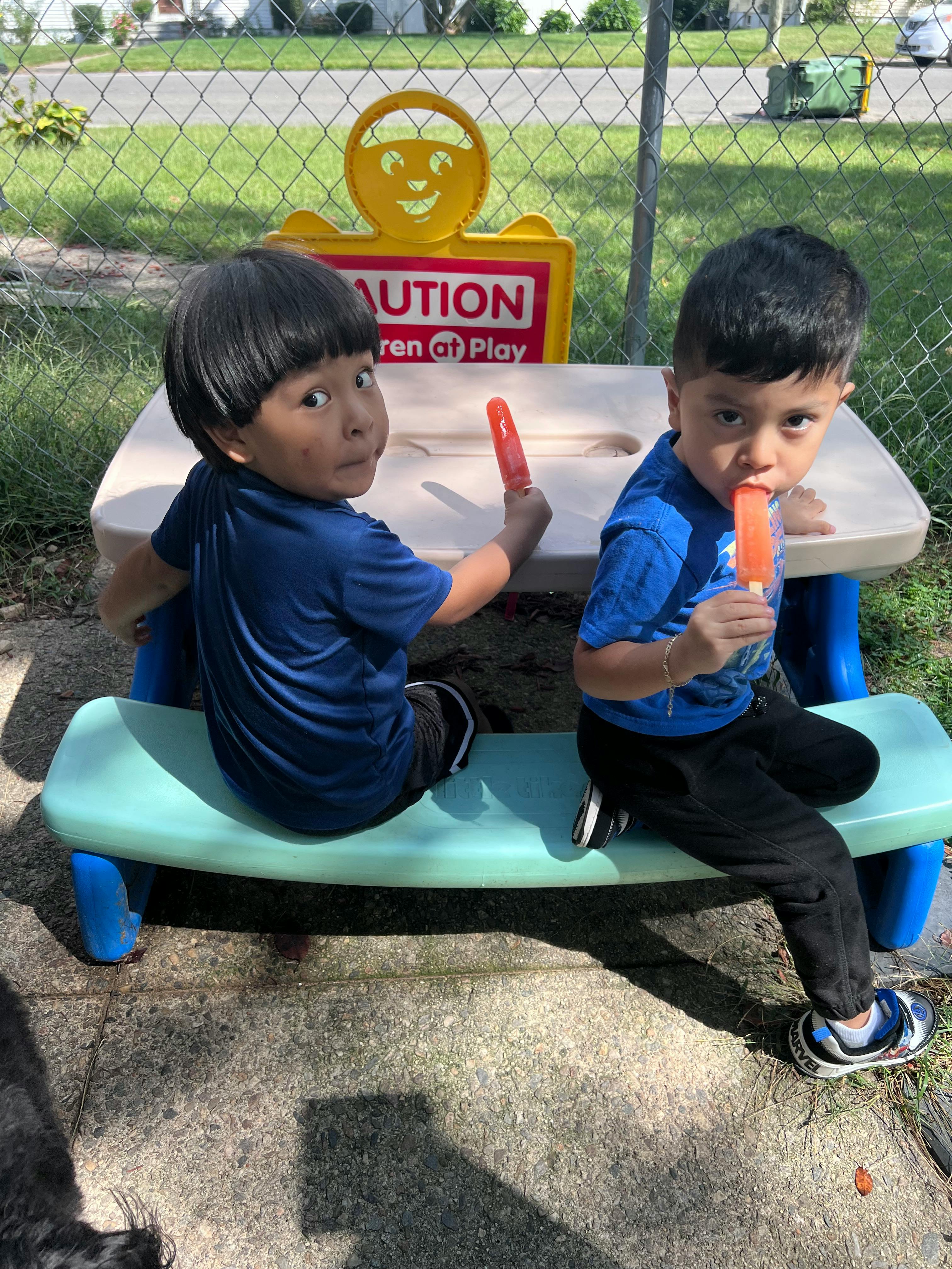Two children eating popsicles at a small table with a "Caution Children at Play" sign