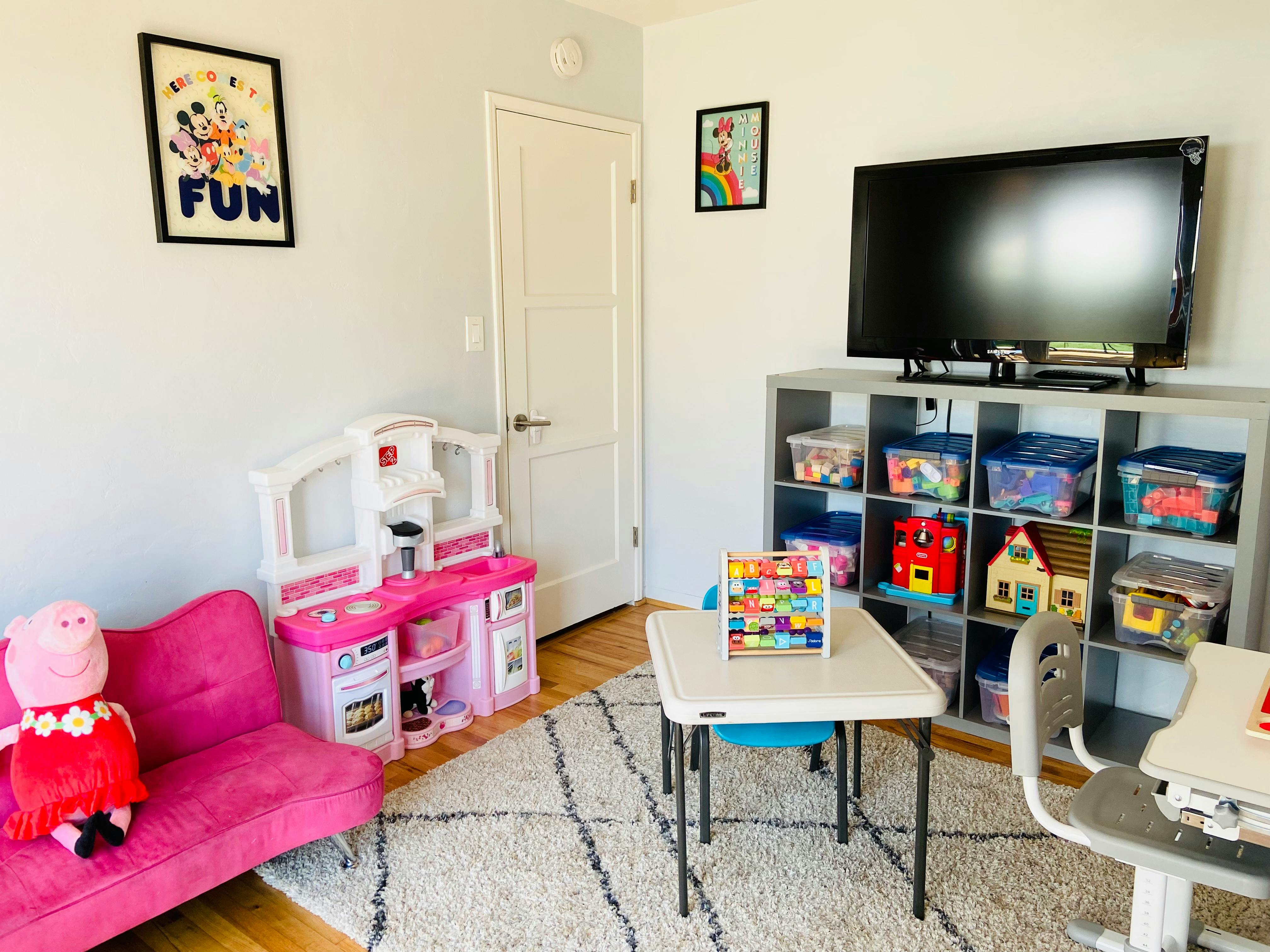 Playroom with a pink toy kitchen, small table, and TV on a storage unit