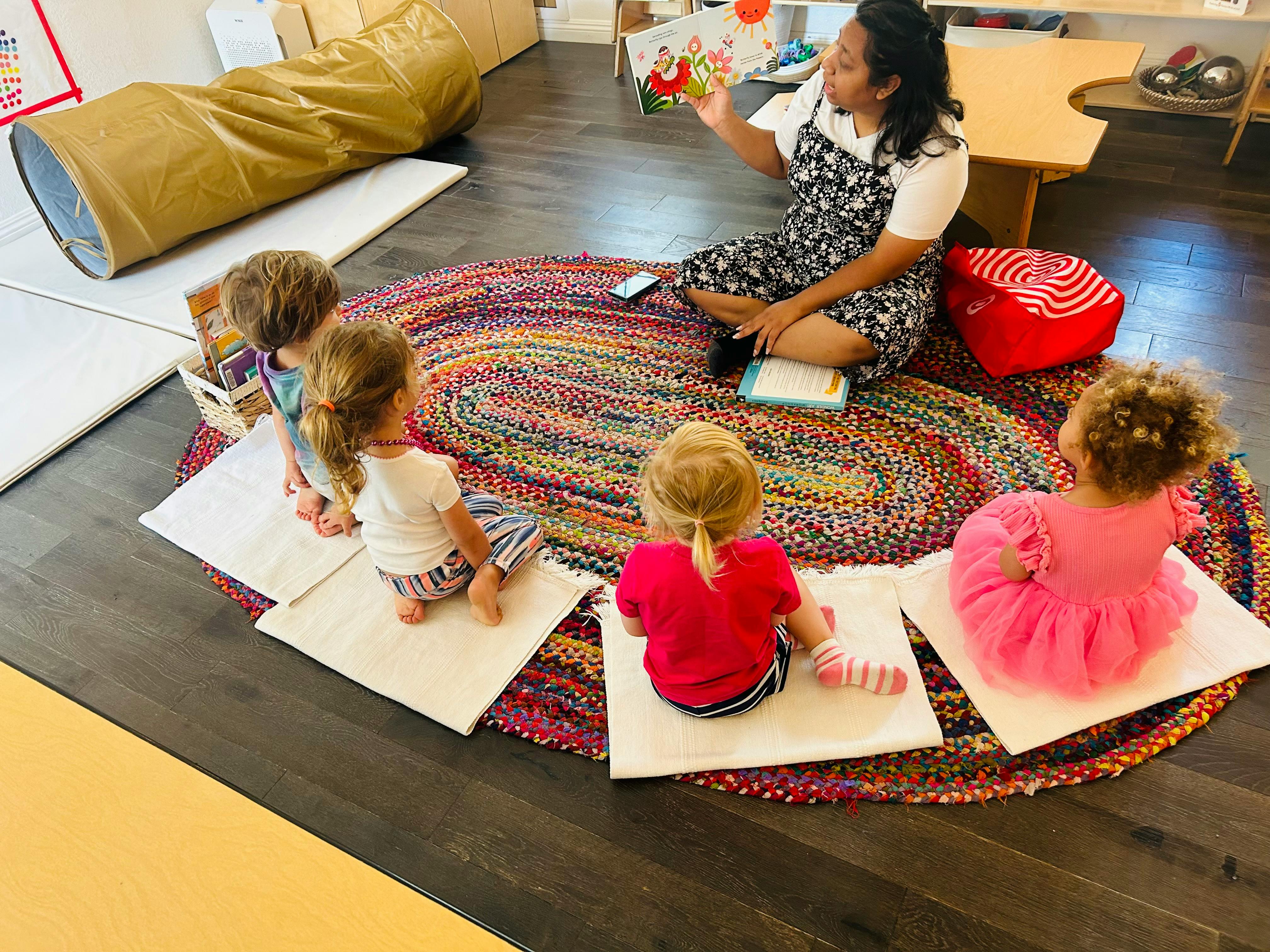 Person reading a book to four children sitting on a rug