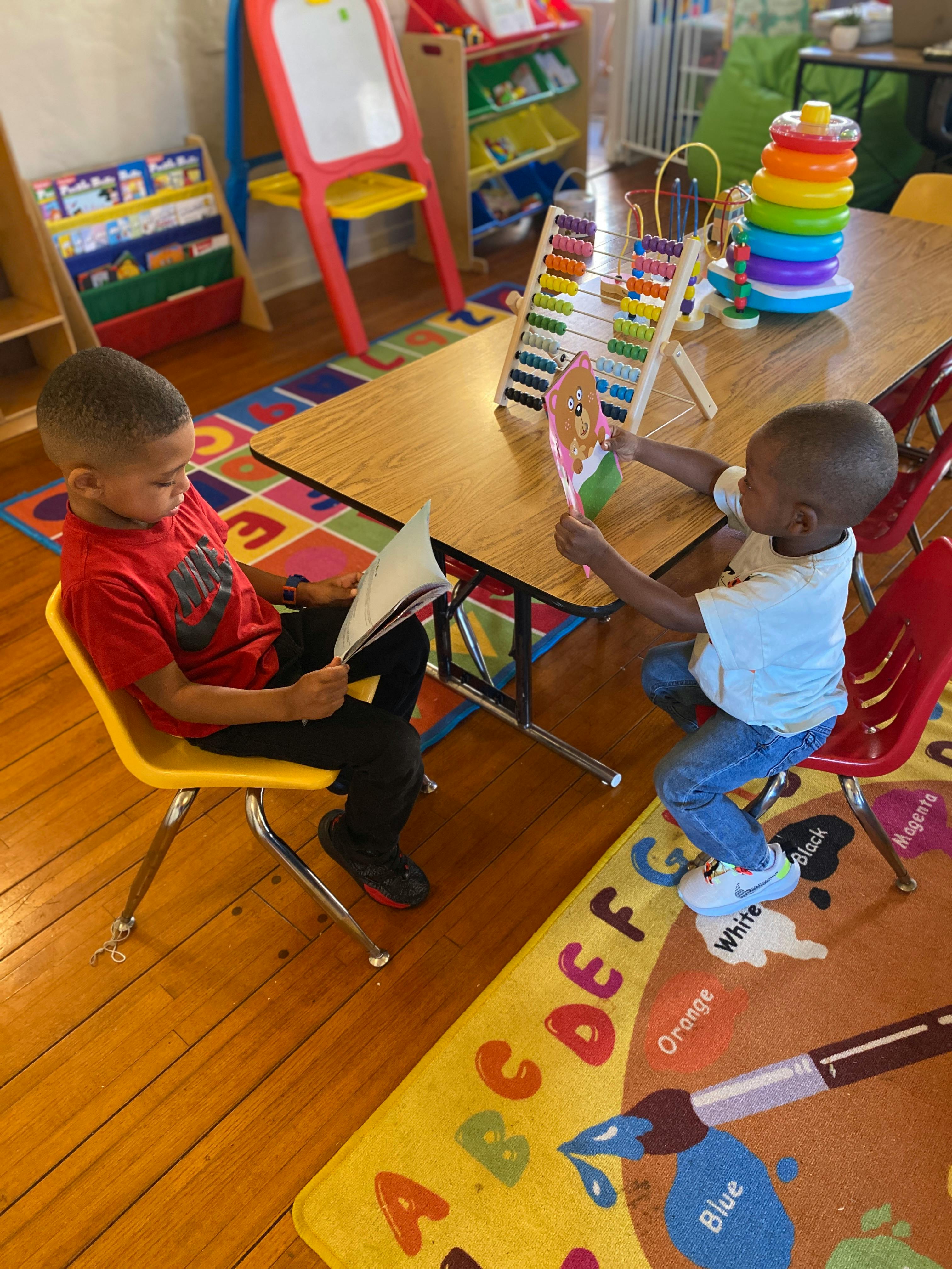 Two children sitting at a table, one reading a book, the other holding a toy
