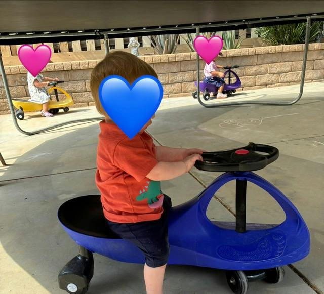 Children riding on plastic swing cars outdoors under a trampoline