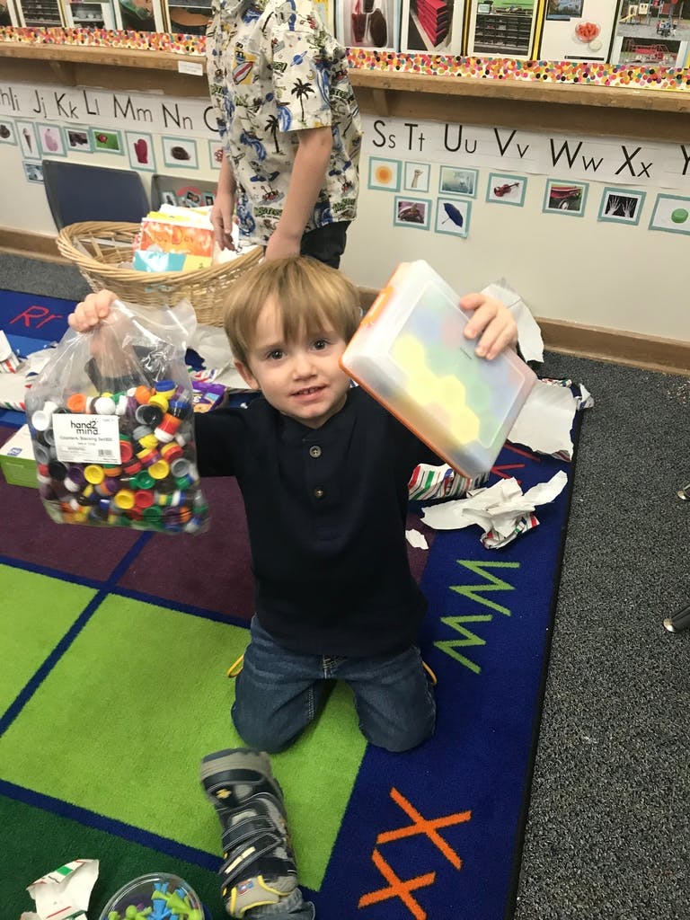 Child kneeling on a rug holding a bag of colorful bottle caps and a container