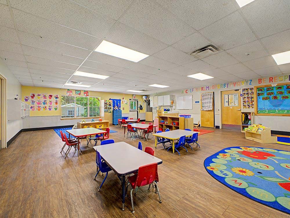 Empty classroom with small tables and chairs, colorful rugs, and artwork on walls