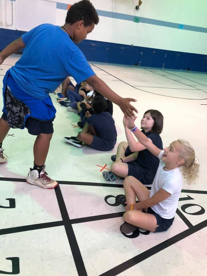 Teen reaching out to high-five a child sitting on the floor in a gym