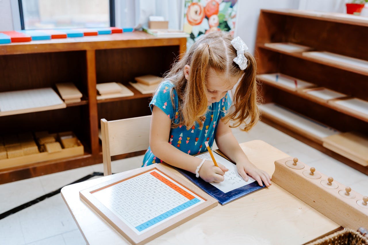 Child writing on paper with a pencil at a table in a classroom