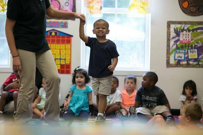 Adult holding a child's hand in a classroom with other children sitting