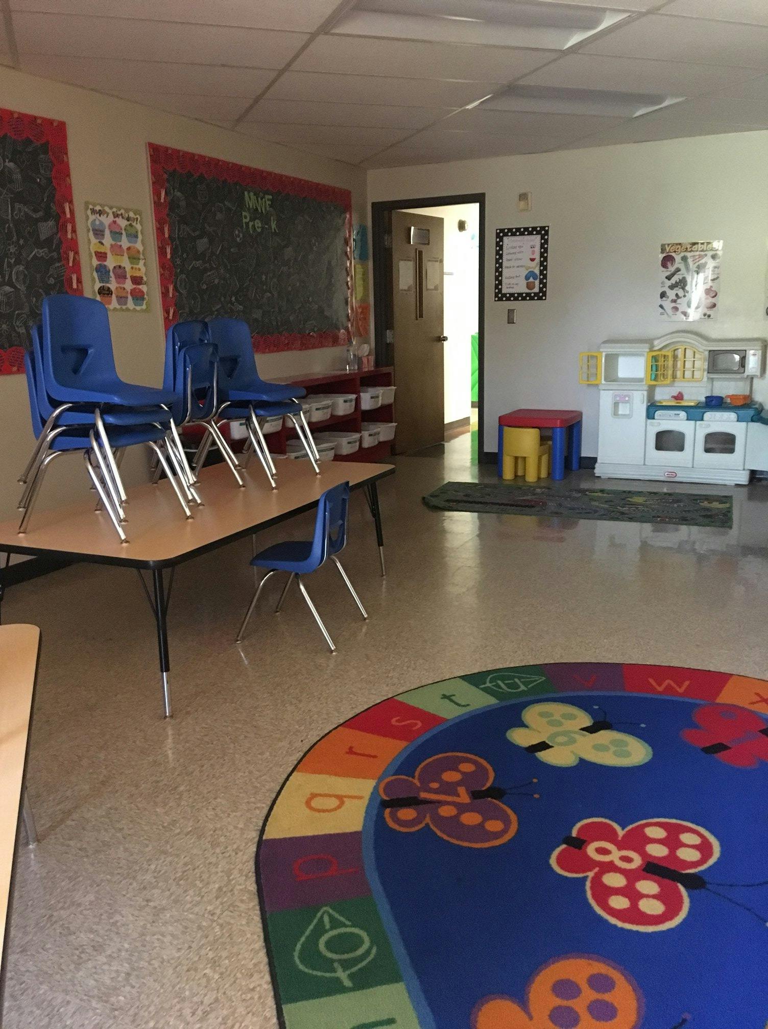 Classroom with stacked chairs on tables, play kitchen, and colorful rug